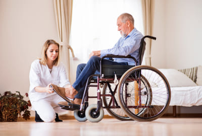 Health visitor or a nurse and a senior men in a wheelchair during home visit. A nurse helping a senior men to put a slipper on.