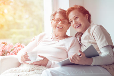 Young caregiver and senior women laughing together while sitting on sofa